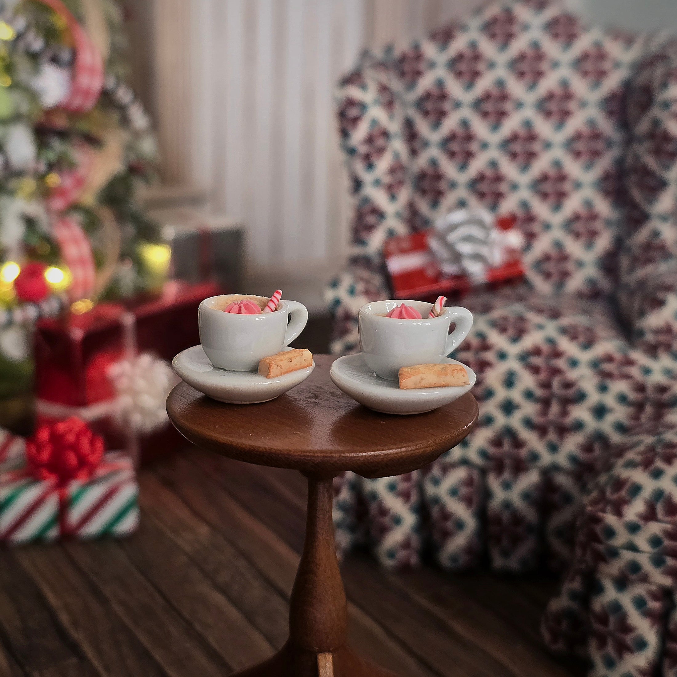 Two miniature cups with saucers on a small wooden table in a cozy room with a Christmas tree and patterned chair.