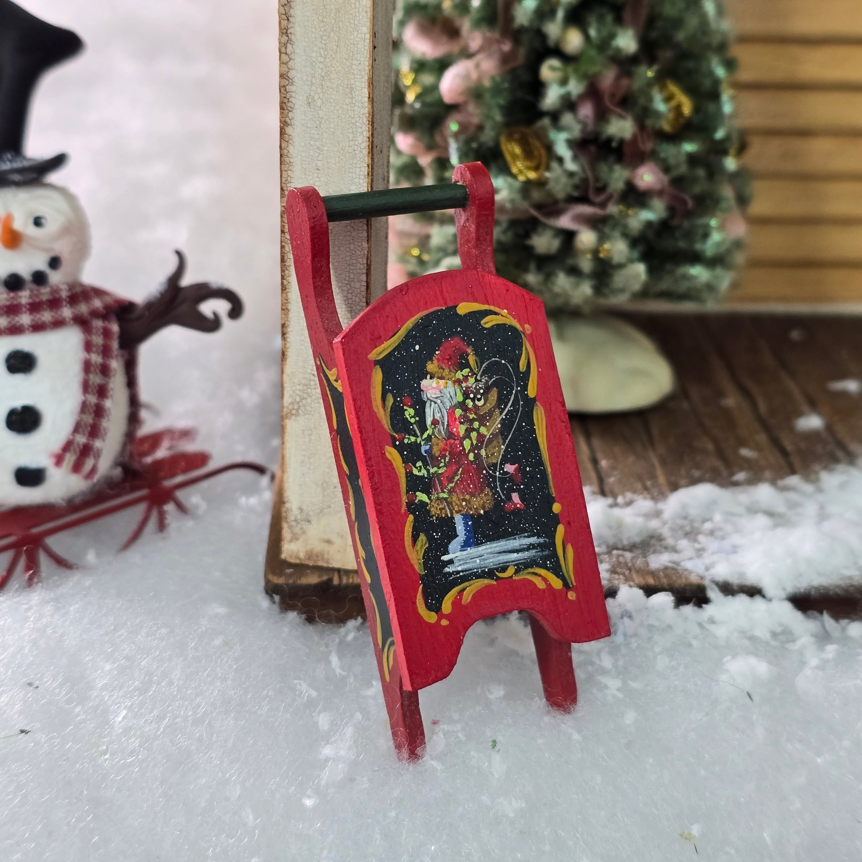 Decorative red sled with a Santa Claus design on a snowy surface with a Christmas scene in the background.