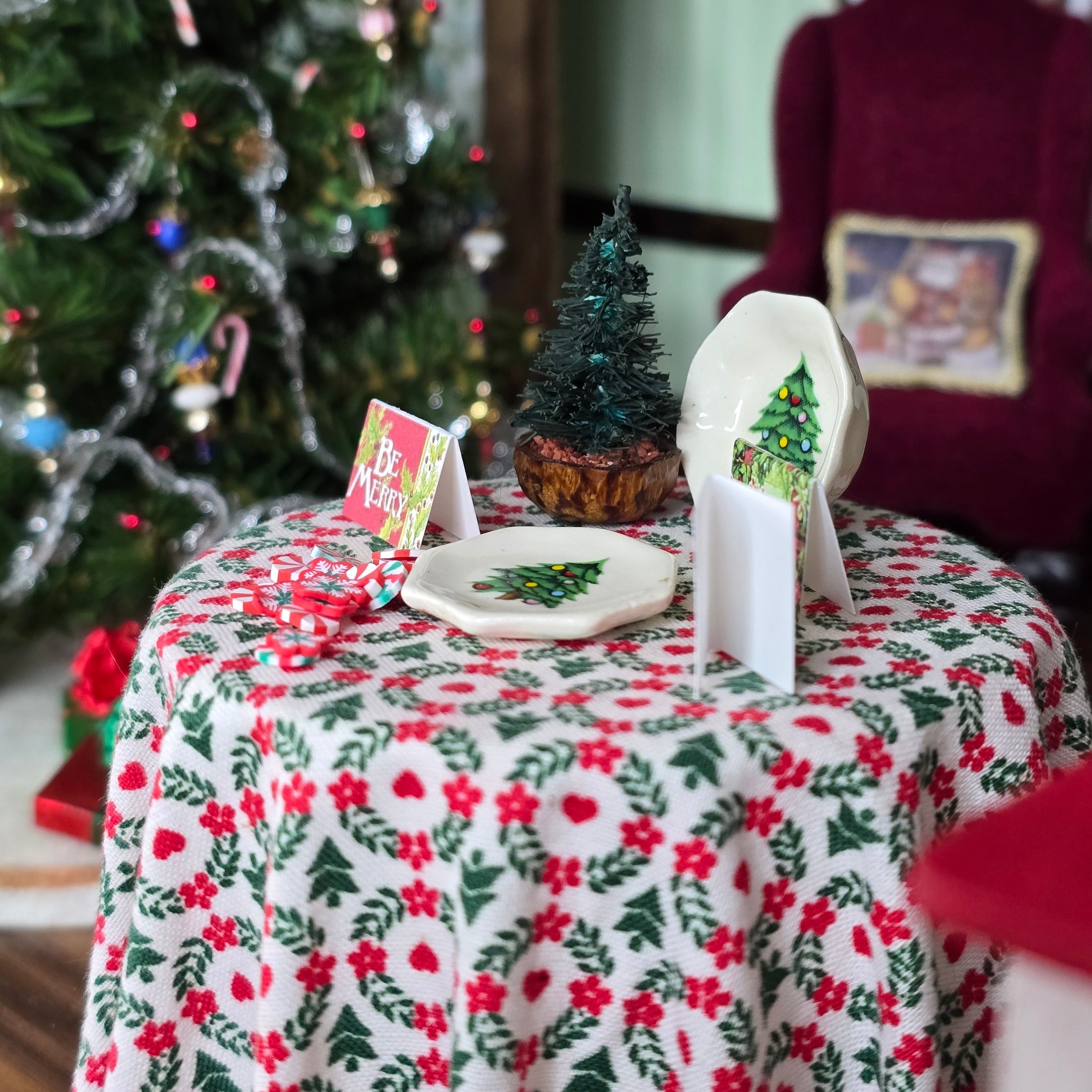 Miniature Christmas table setting with decorative items and a tree in the background.