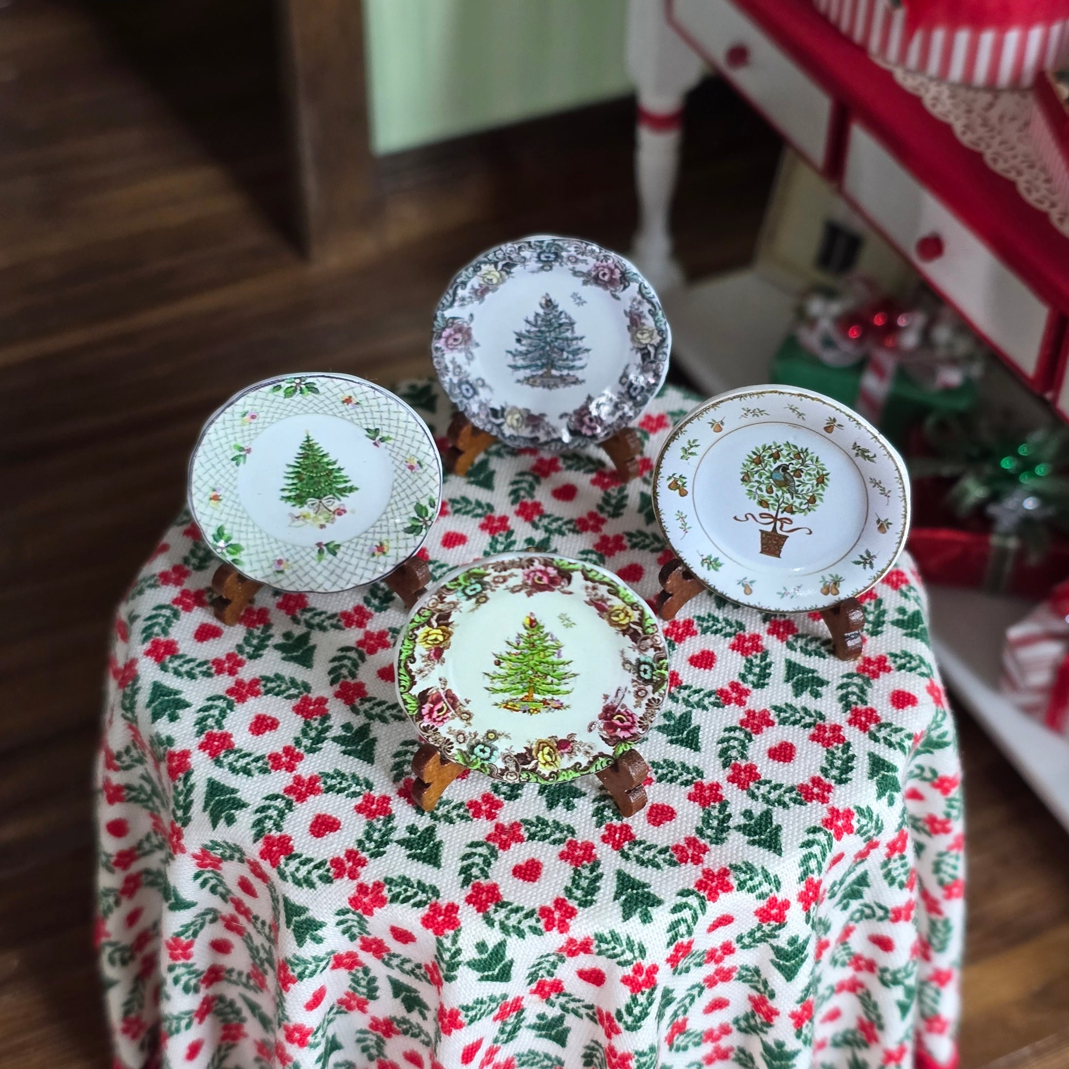 Four decorative plates with Christmas tree designs on a tablecloth with holly pattern.