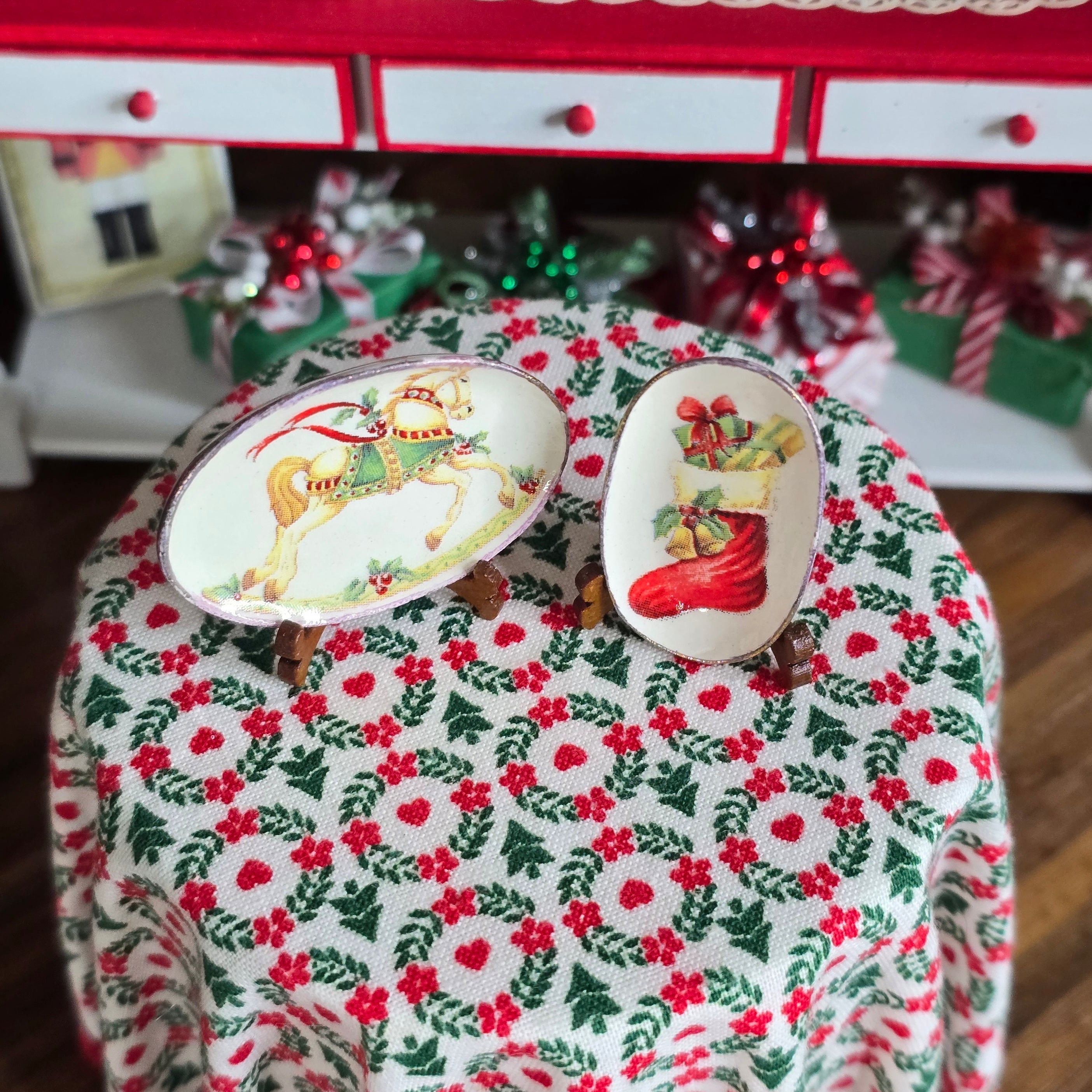 Decorative dollhouse table setting with Christmas-themed plates on a patterned tablecloth.