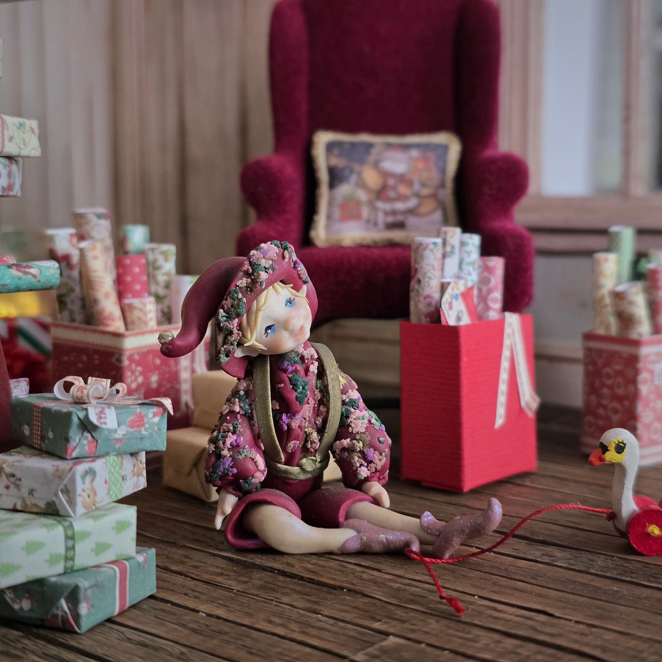 Hand-Sculpted Clay Elf Boy in Raspberry Pink in a floral outfit surrounded by gift boxes on a wooden floor.