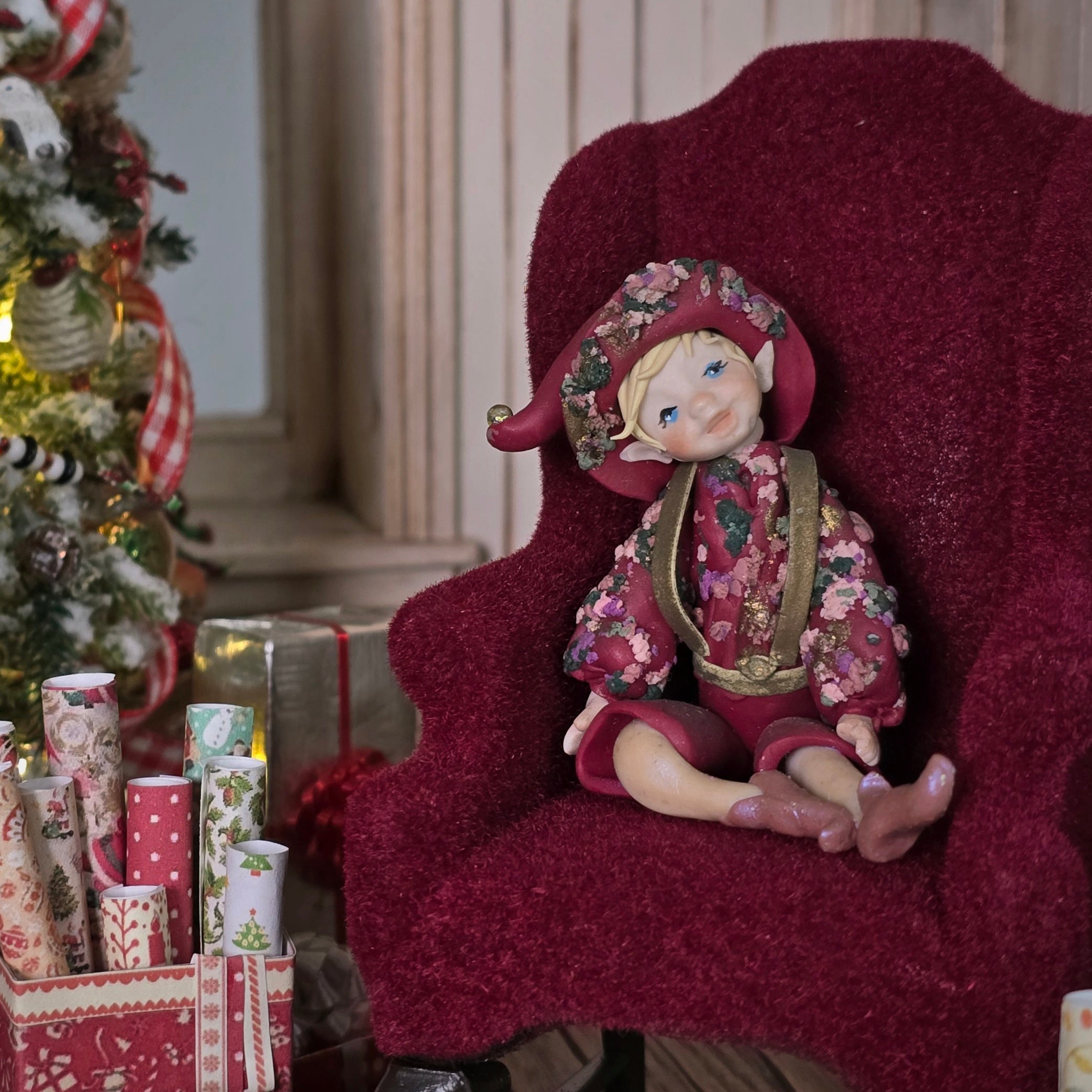 Hand-Sculpted Clay Elf Boy in Raspberry Pinkl in a floral outfit sitting on a red chair with Christmas decorations in the background.