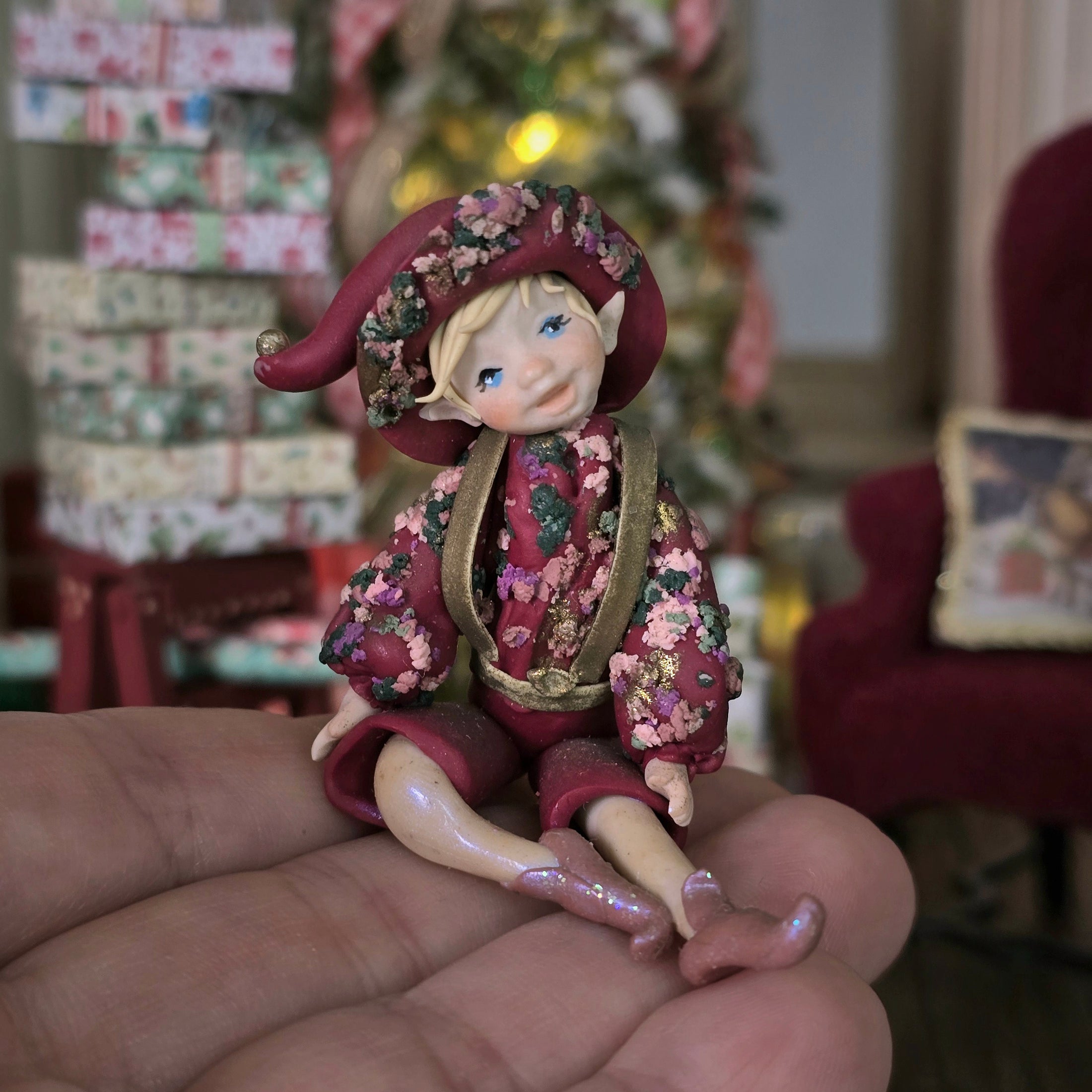 Hand-Sculpted Clay Elf Boy in Raspberry Pink floral suir, held in a hand with a decorated room in the background