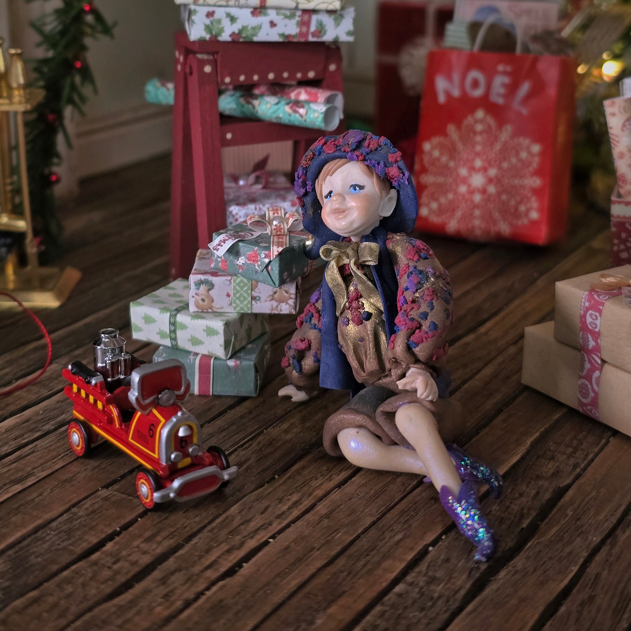 Sculpted clay elf boy doll wearing a purple outfit with a toy fire truck and Christmas presents on a wooden surface