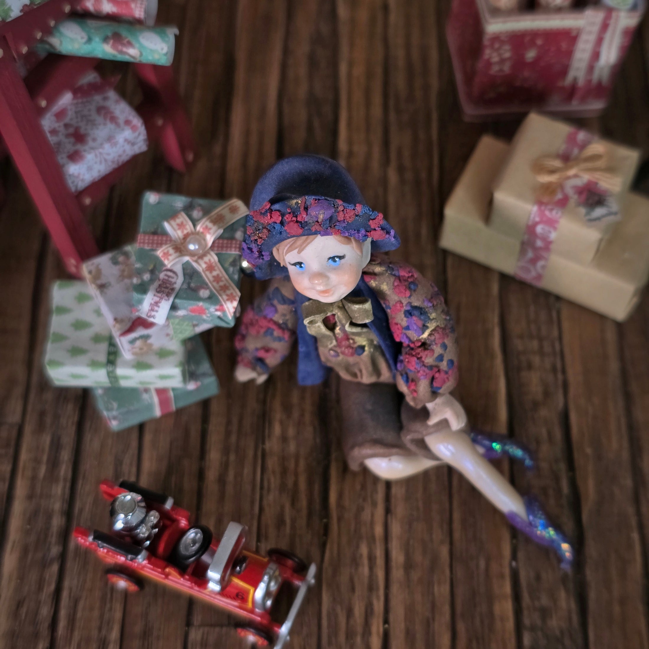 top view of a Sculpted clay elf boy doll wearing a purple outfit with floral hat and coat surrounded by small presents on a wooden surface