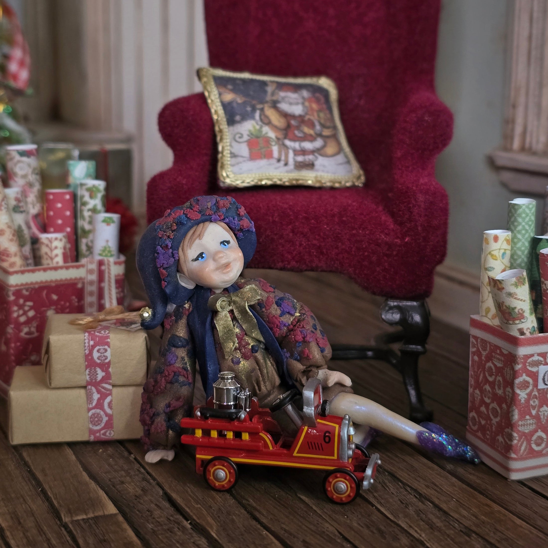Sculpted clay elf boy doll wearing a purple outfit with a toy fire truck in a festive setting with presents and a red chair.