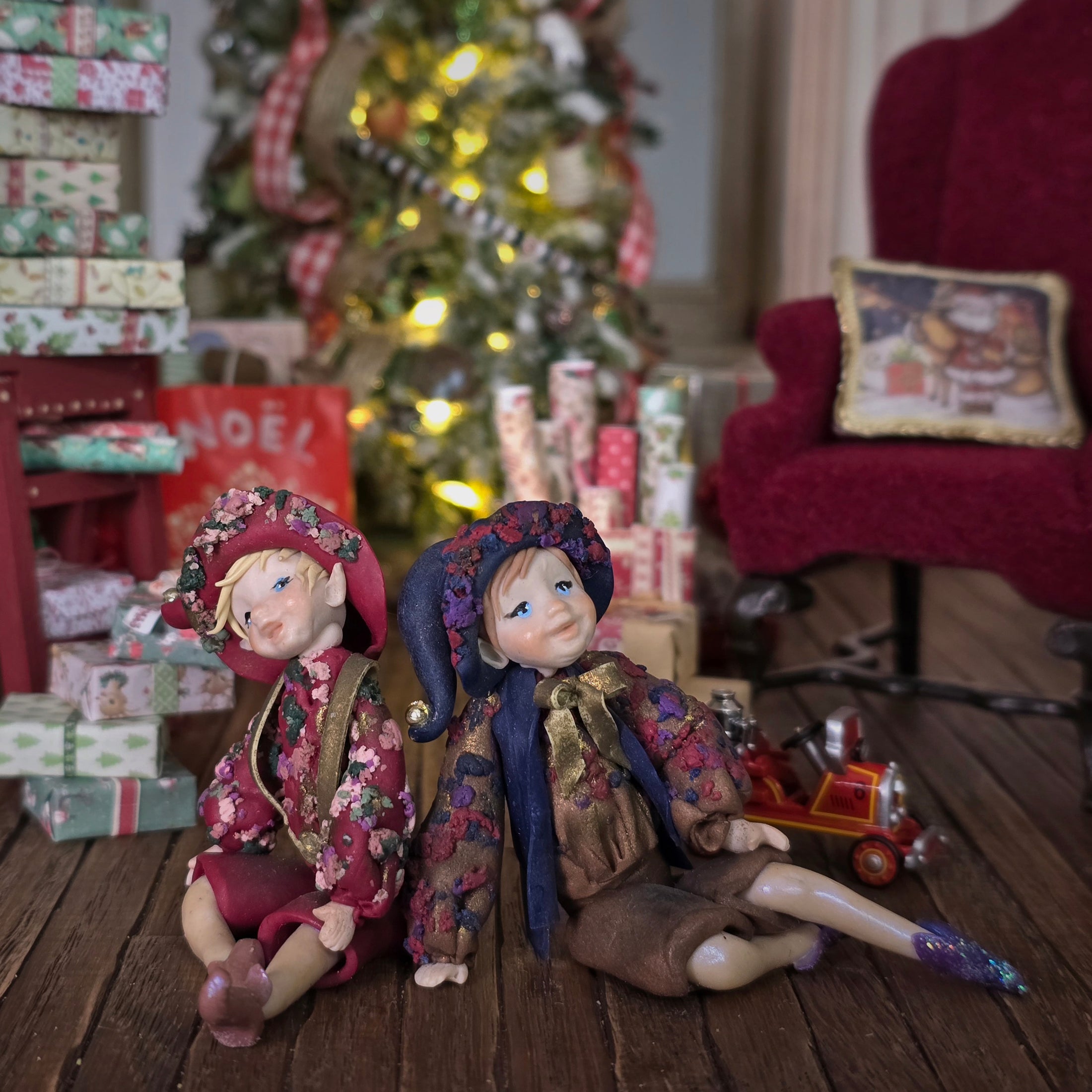 Two Sculpted clay elf boy doll in festive attire sitting on a wooden floor with a decorated Christmas tree and presents in the background.