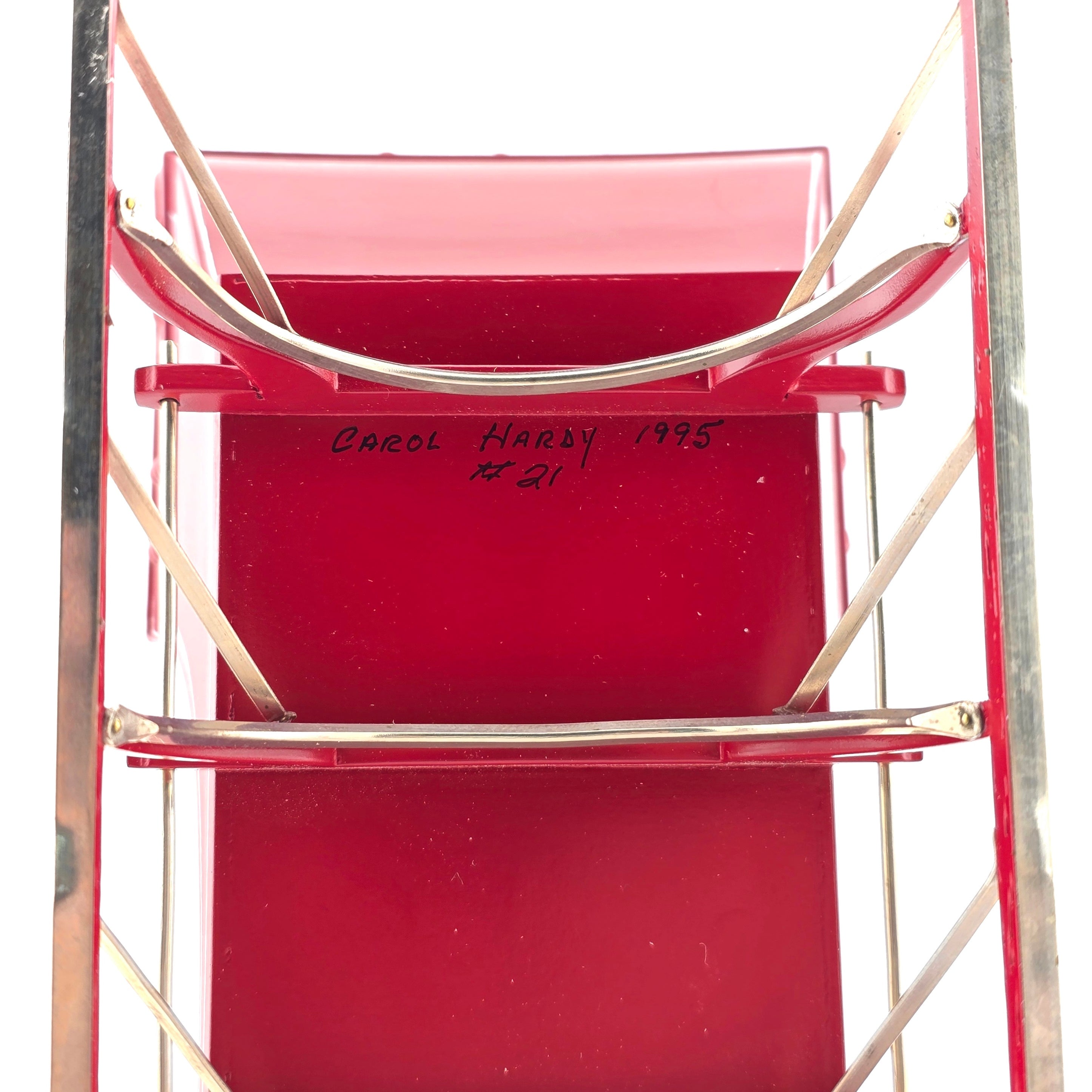 Red metal chair with 'Carol Hardy 1995 #21' written on it, against a white background.