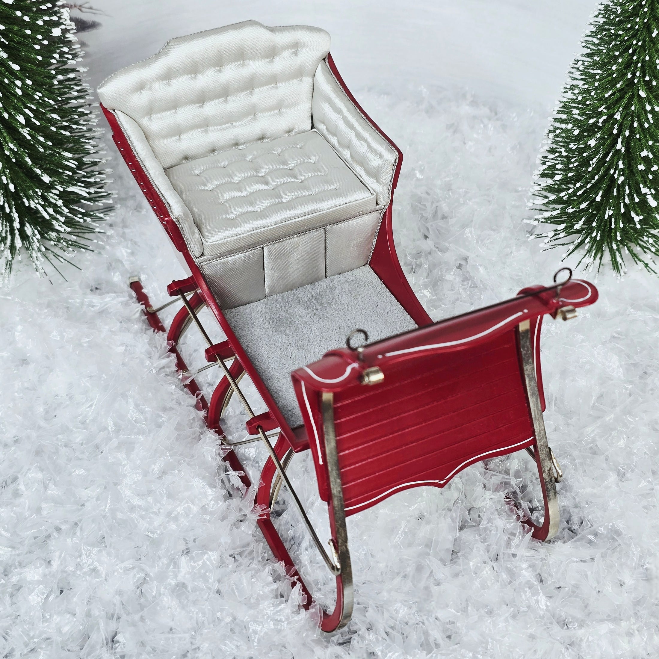 Red sleigh with white seat on a snowy ground with Christmas trees.