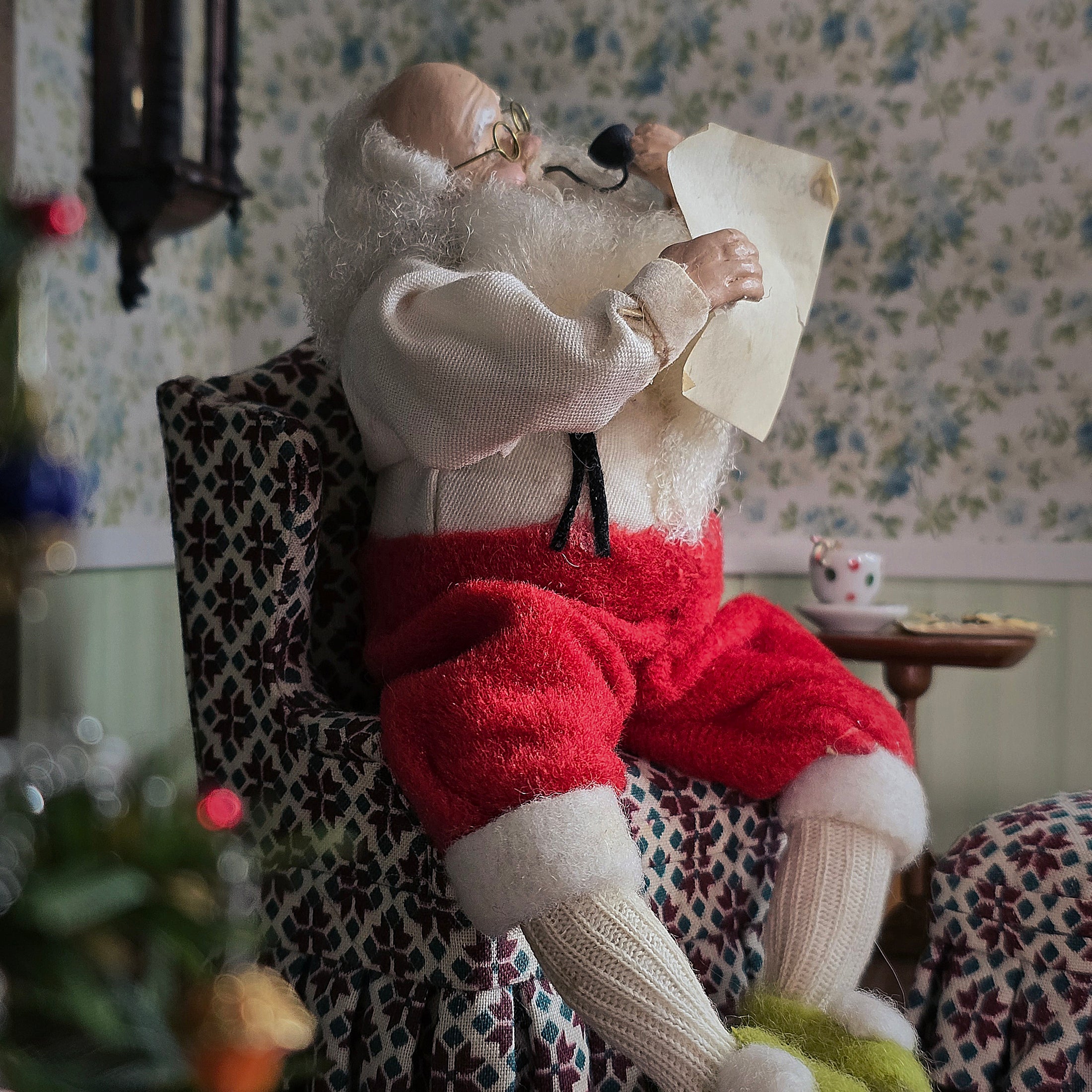 Santa Claus sitting in a chair reading a letter in a cozy room with floral wallpaper.