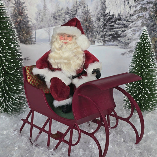 Santa Claus dollhouse doll sitting in a sleigh in a snowy landscape with trees.