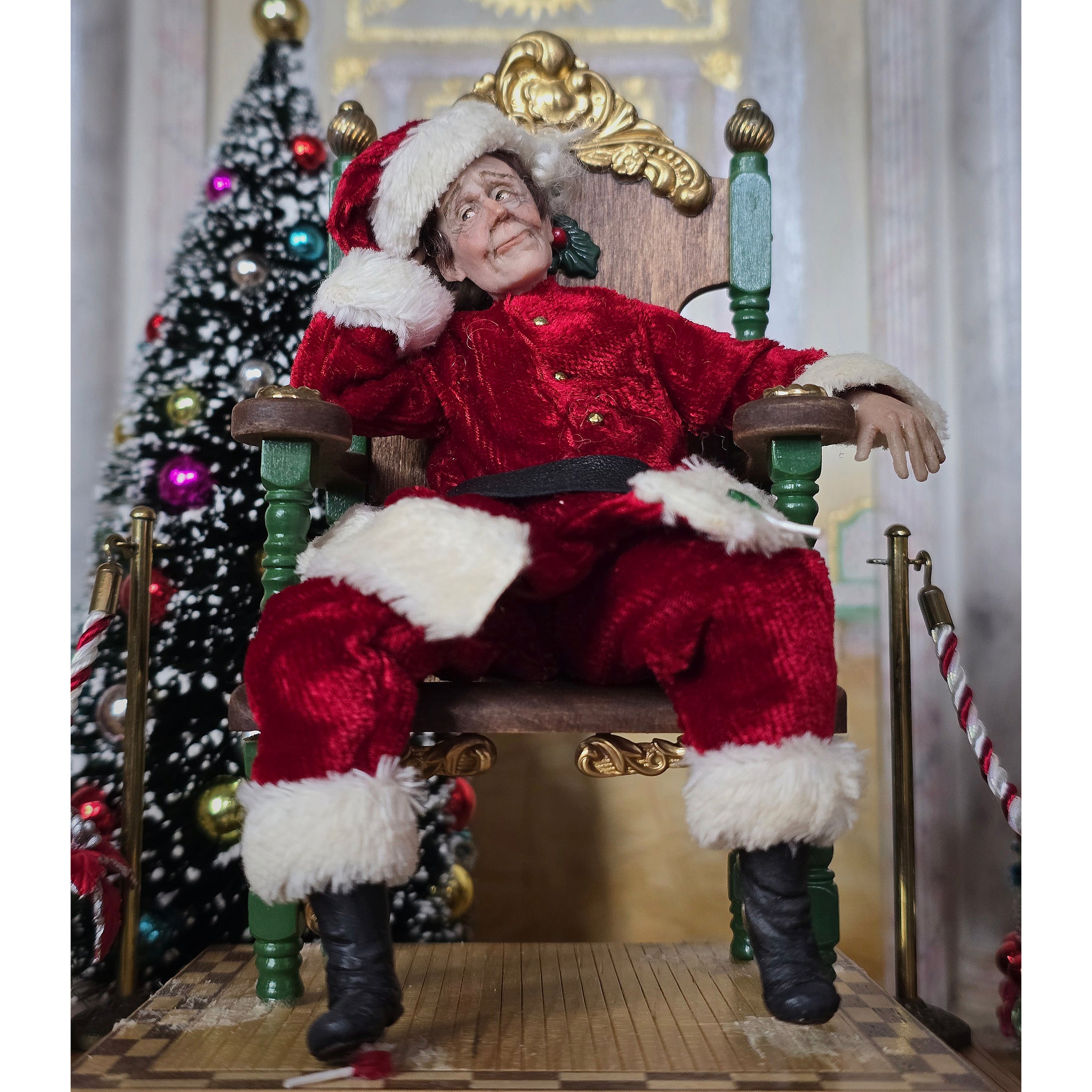 Santa dollhouse doll sitting in a throne in a festive setting with a decorated Christmas tree.