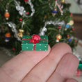 Load image into Gallery viewer, Miniature green gift box with a red ribbon held in front of a decorated Christmas tree.
