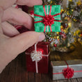 Load image into Gallery viewer, Hand holding a small green gift box with a red bow in front of a Christmas tree.
