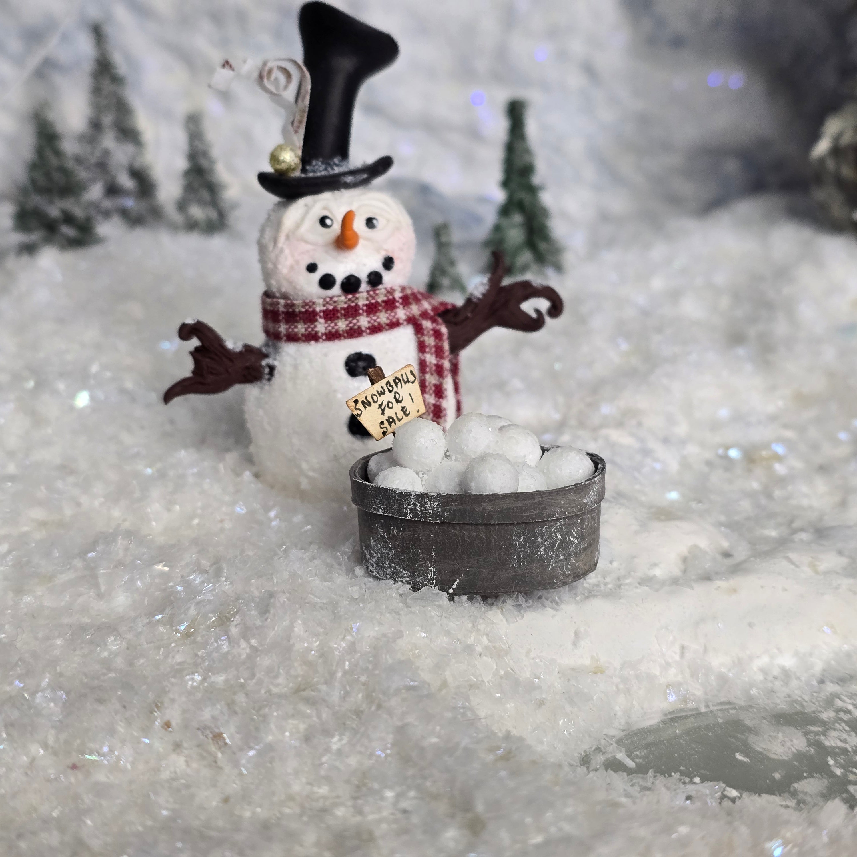 Decorative snowman miniature with a top hat and scarf, standing near a small bucker on a snowy background.