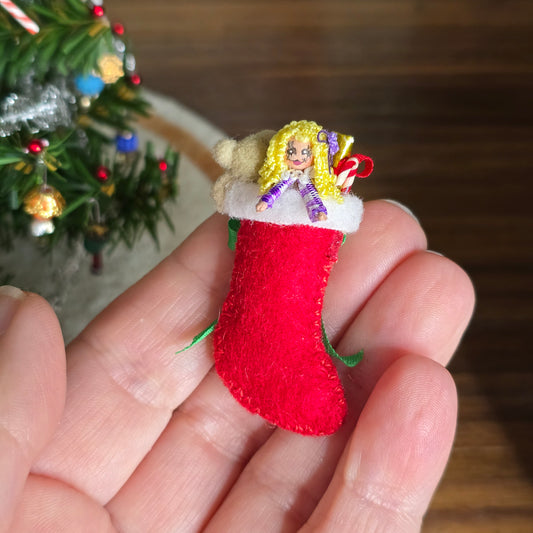 Miniature red Christmas stocking with a blonde dolly held in a hand, with a blurred Christmas tree in the background.