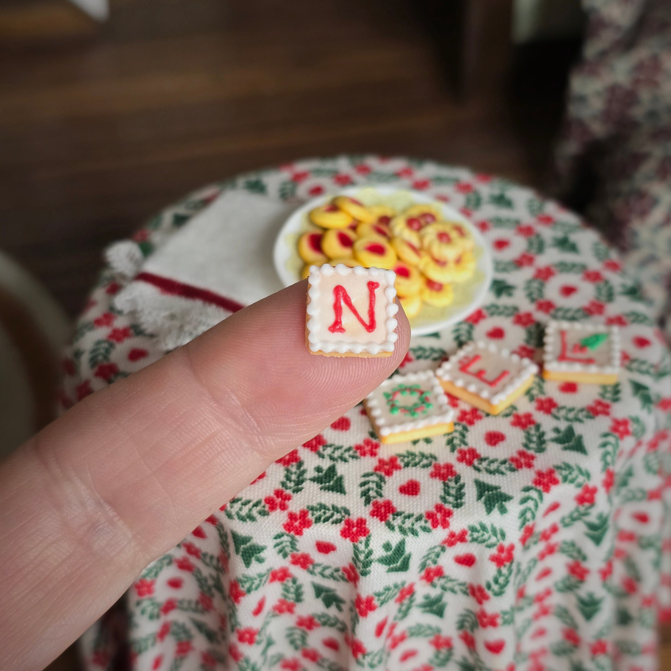 Hand holding a small square cookie with 'N' on it, with a decorative tablecloth in the background.