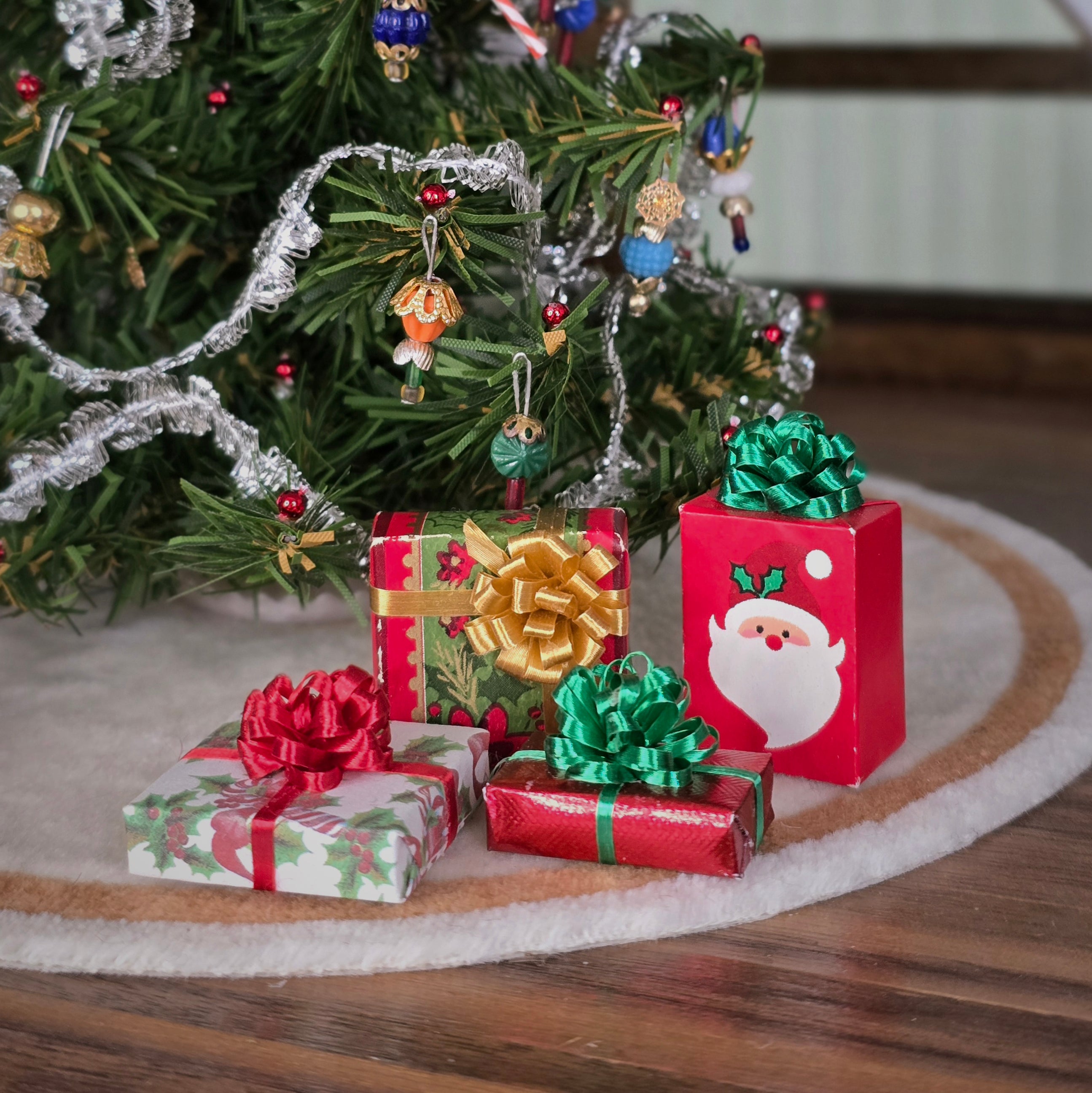 Miniature Christmas presents and ornaments on a wooden surface with a blurred Christmas tree in the background.