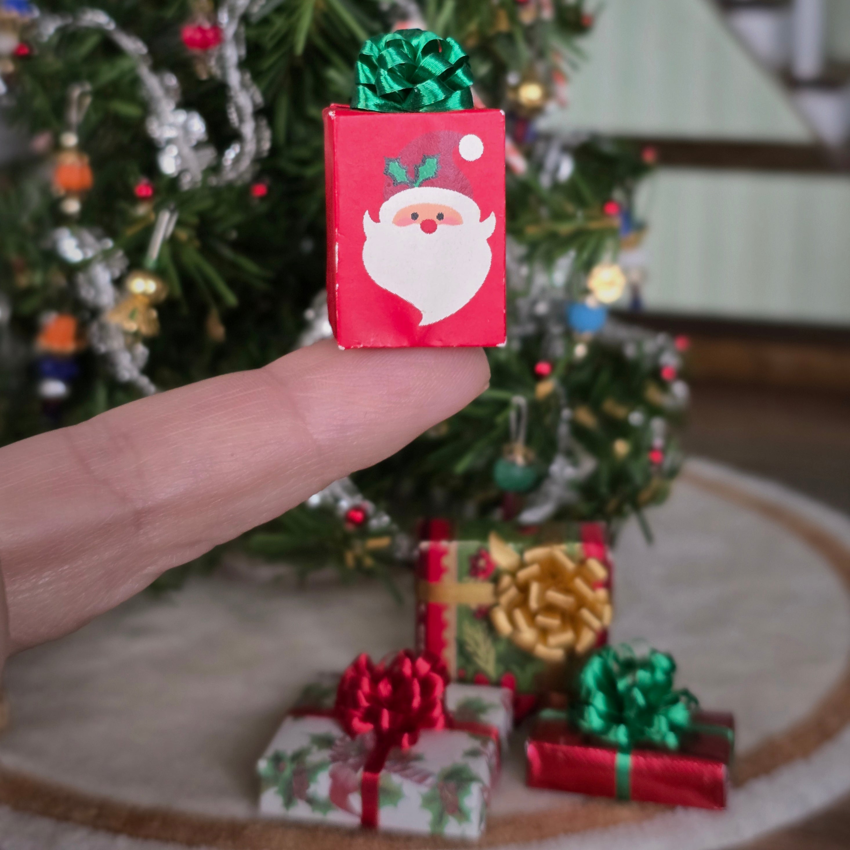 Miniature Christmas gift box with Santa Claus design held by a hand in front of a decorated Christmas tree.