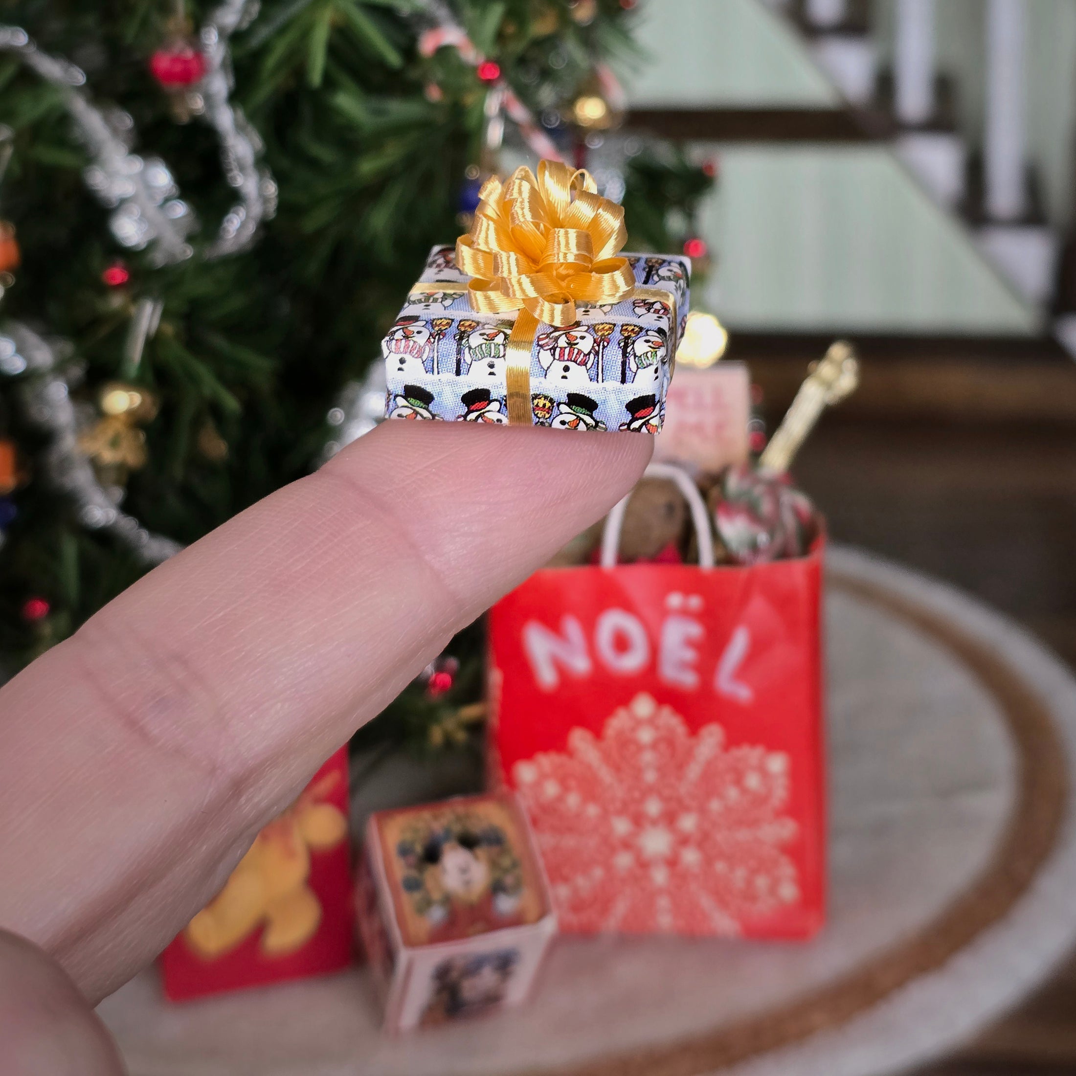 Hand holding a small decorative box with a gold bow in front of a Christmas tree and red 'NOËL' bag.