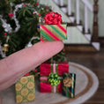 Load image into Gallery viewer, Dollhouse Miniature gift box with a red ribbon held by a finger in front of other small Christmas presents.
