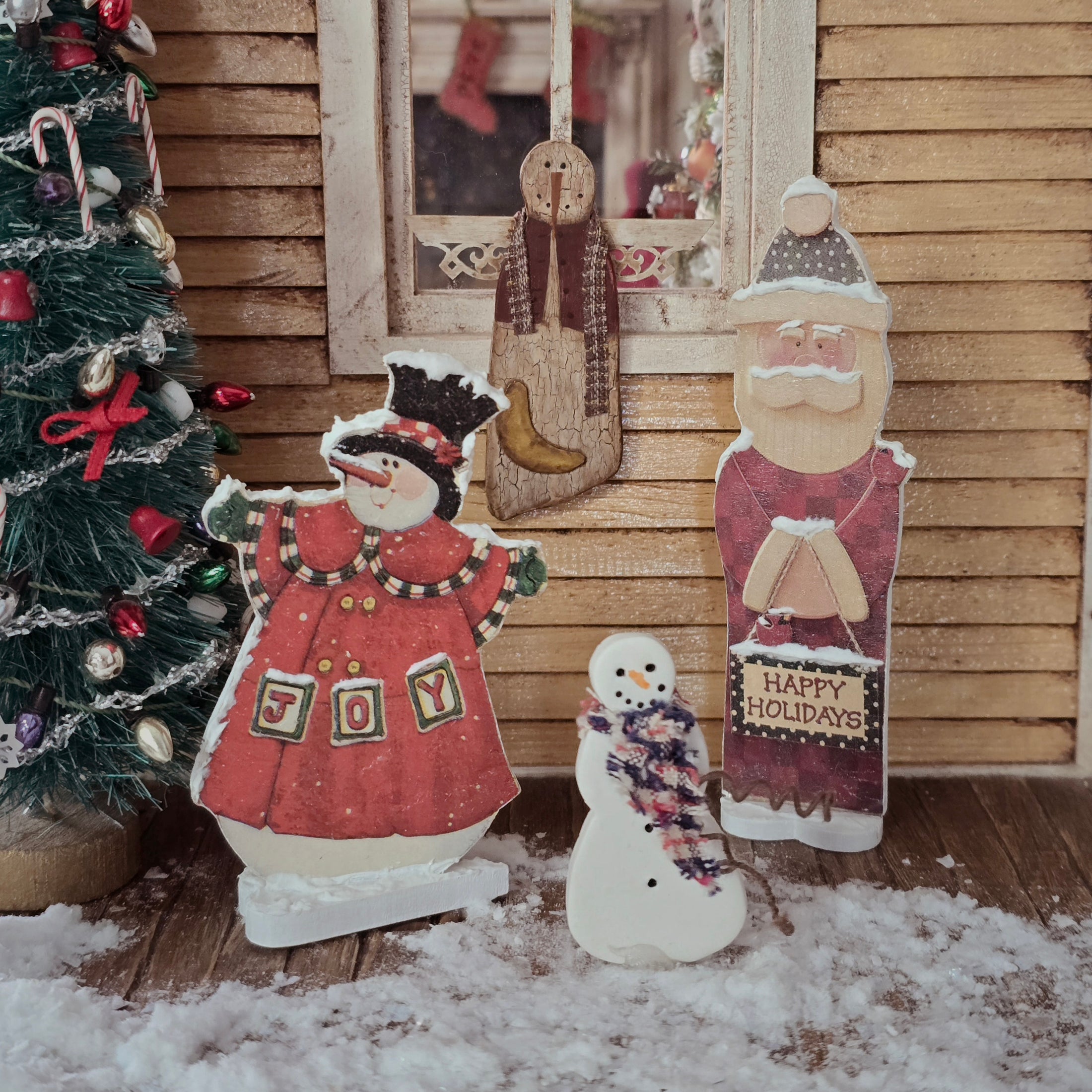 Dollhouse miniature Wooden Christmas decorations including a snowman, Santa, and a snowman with 'Joy' on its coat, in front of a decorated Christmas tree.