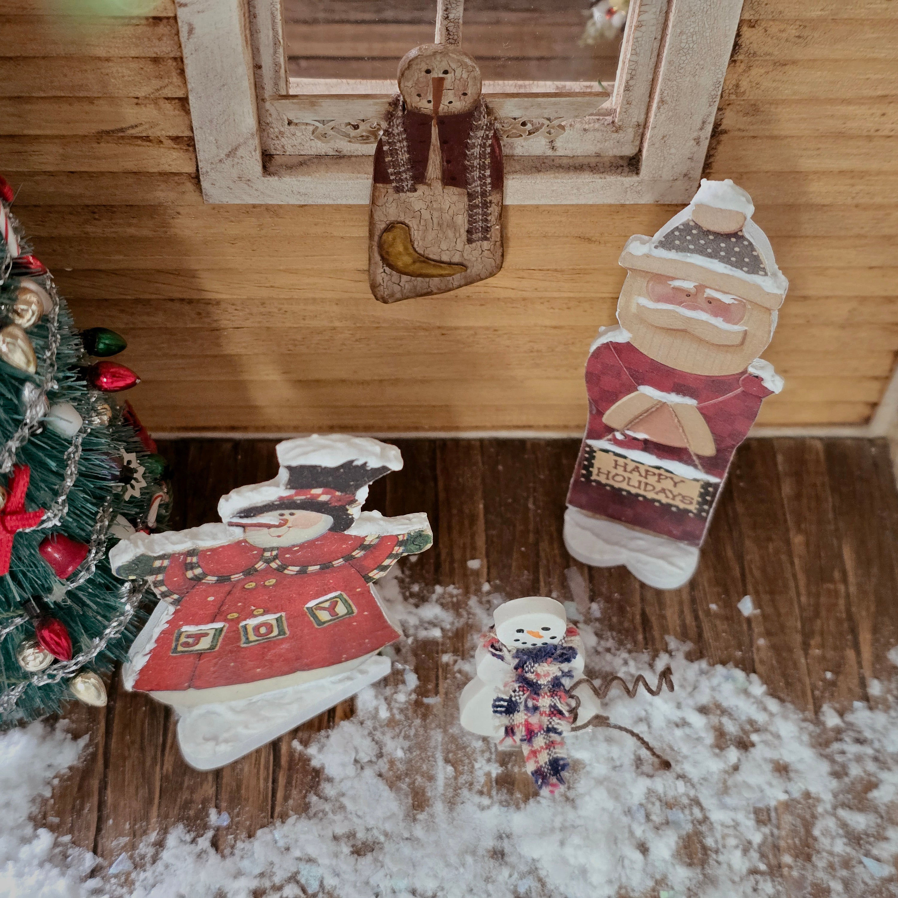 Dollhouse miniature Decorative Christmas figures on a wooden porch with a small tree in the corner.