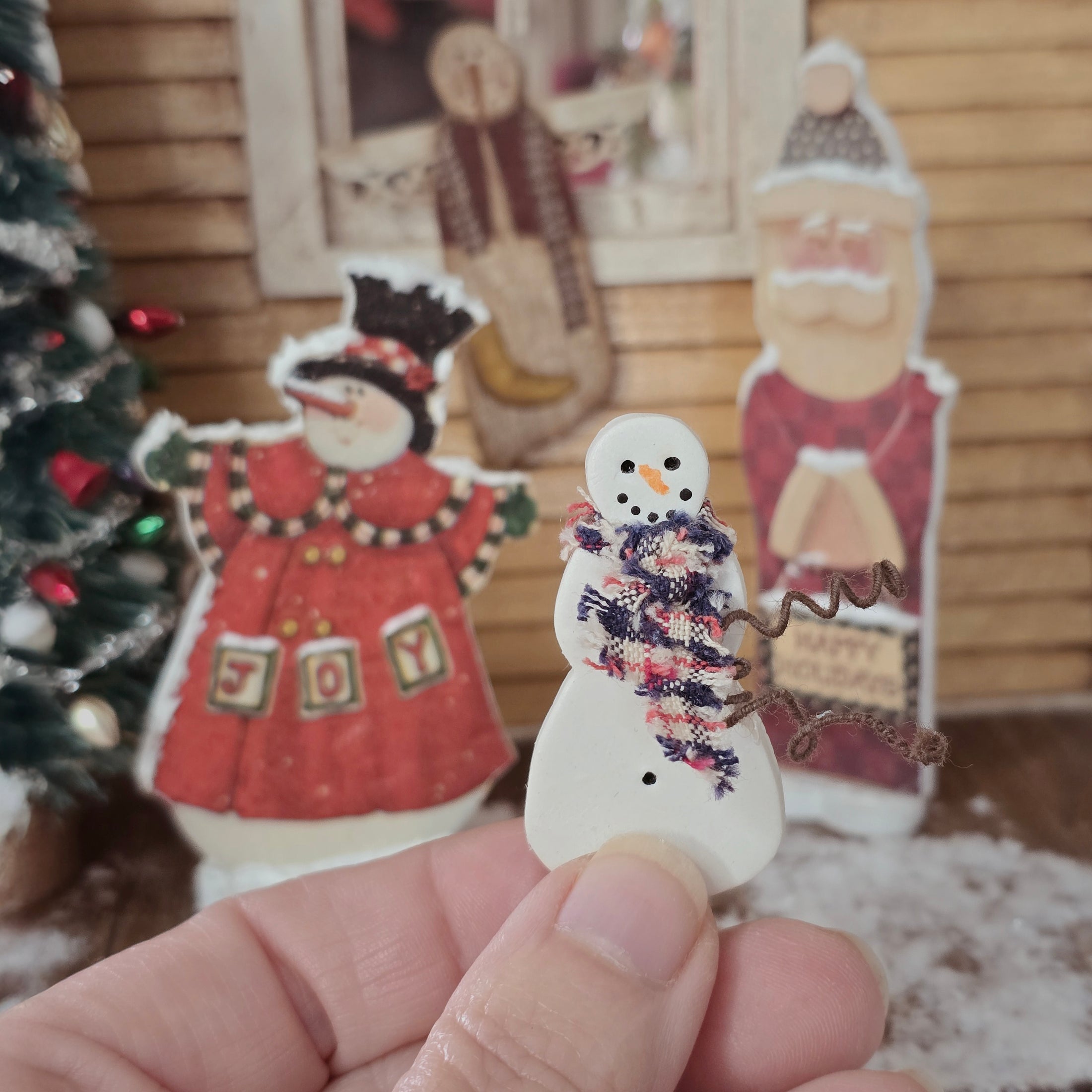 Hand holding a Dollhouse miniature decorative snowman with scarf in front of Christmas decorations.