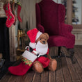 Load image into Gallery viewer, Dollhouse Teddy bear in a Santa outfit sitting on a wooden floor with a red chair and stockings in the background.
