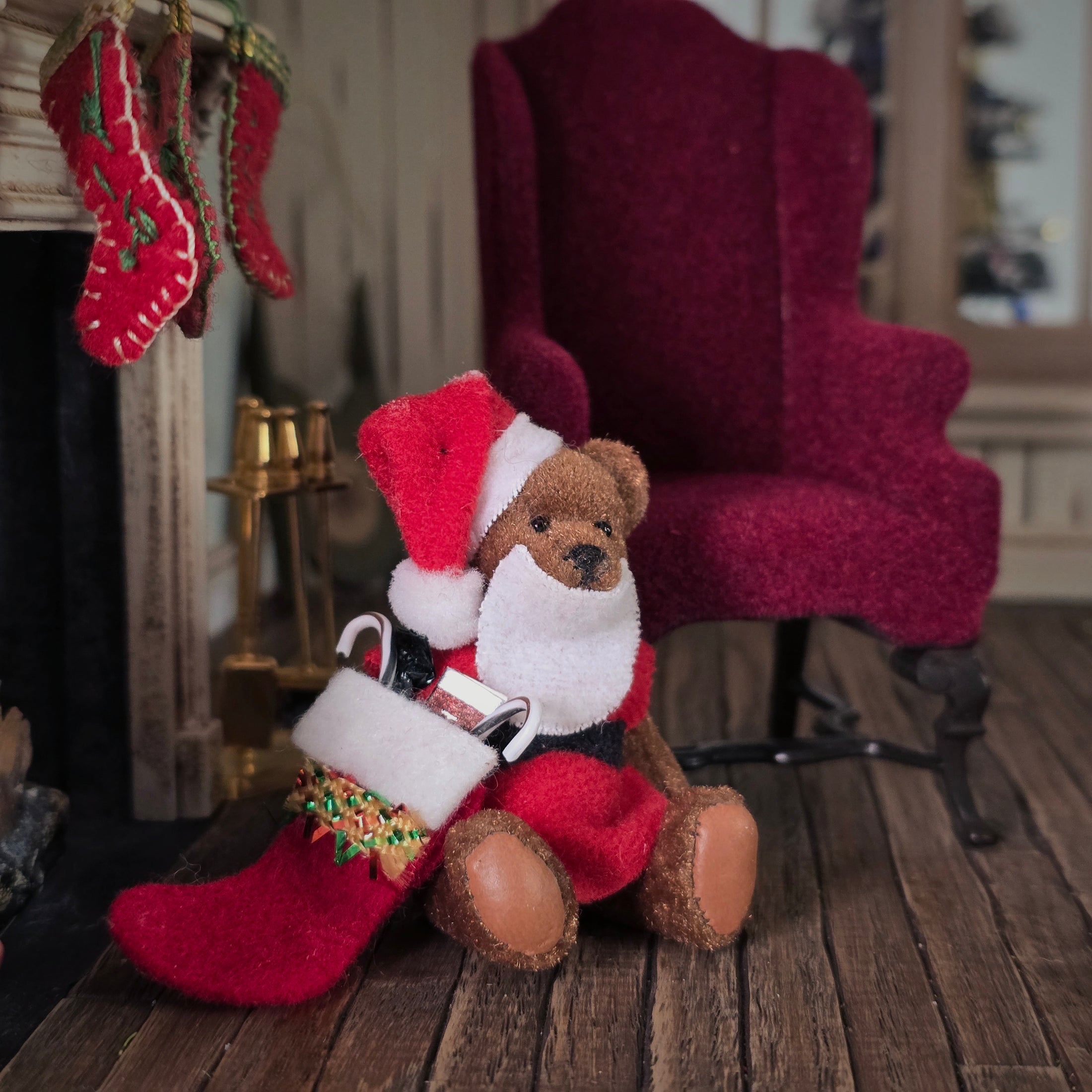 Dollhouse Teddy bear in a Santa outfit sitting on a wooden floor with a red chair and stockings in the background.