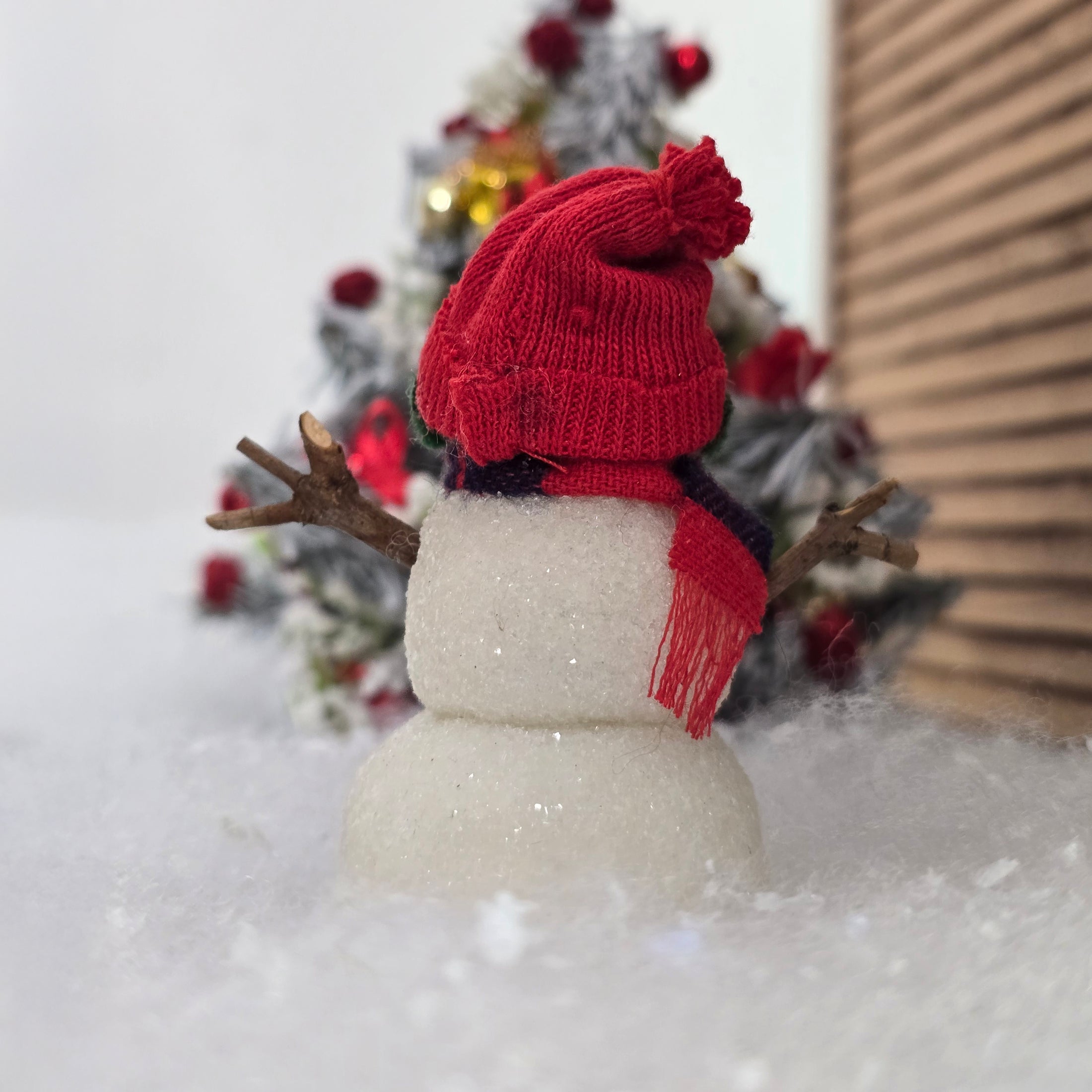Back view of a small snowman with a red hat and scarf in front of a Christmas tree.