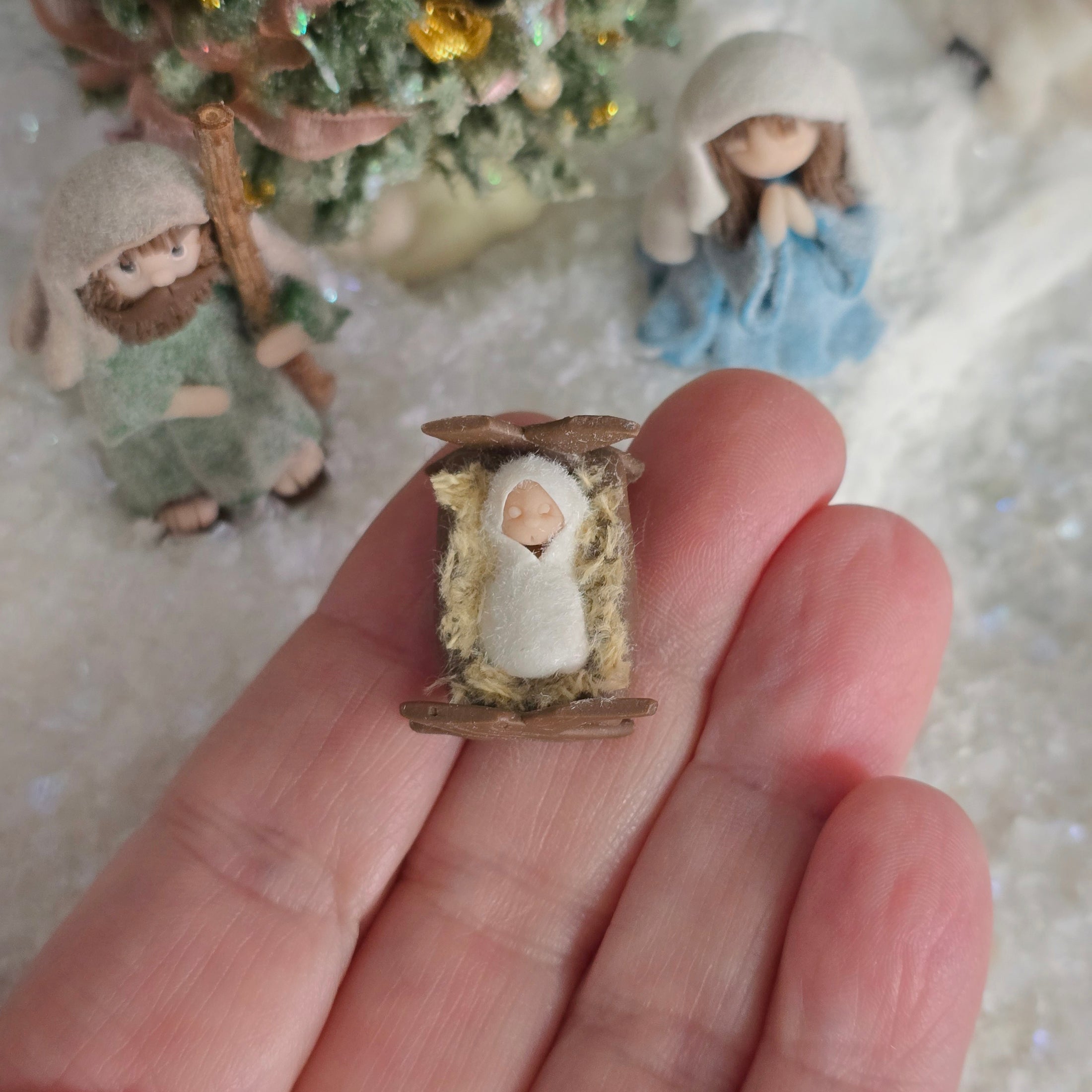 Miniature Bby Jesus in a manger held in a hand with other nativity figures in the background. miniature dollhouse