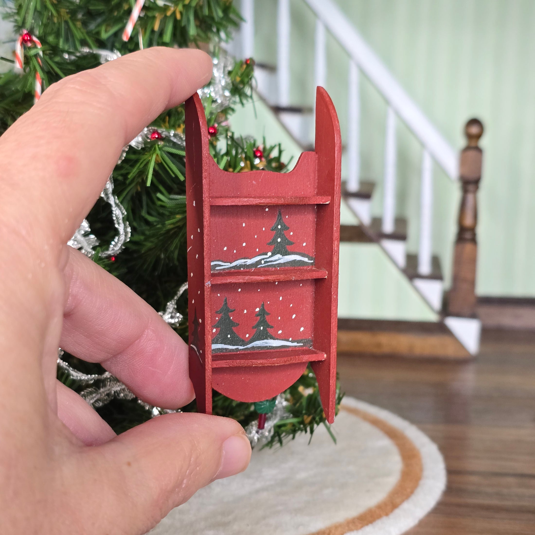 Hand holding a small red sled with Christmas tree design in front of a decorated Christmas tree.
