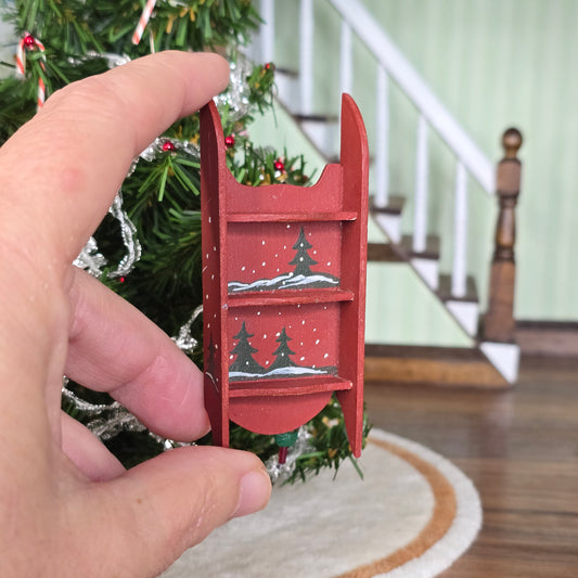 Hand holding a small red sled with Christmas tree design in front of a decorated Christmas tree.