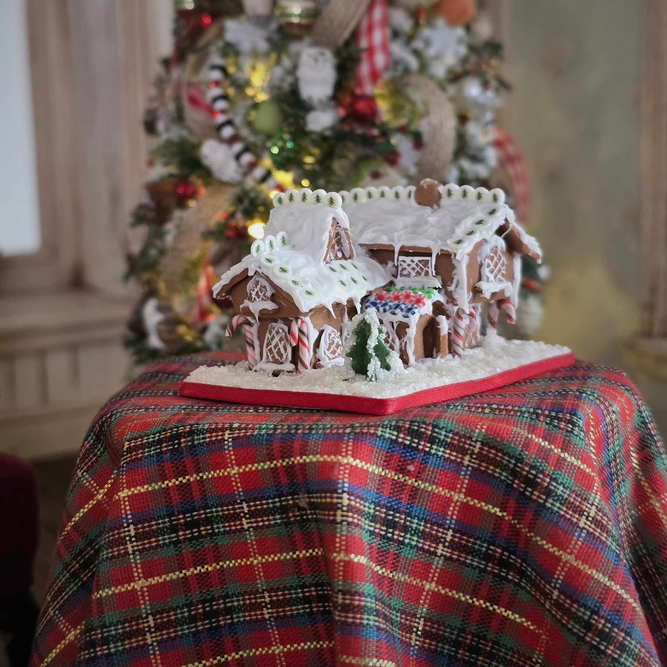 Gingerbread house on a plaid tablecloth with a decorated Christmas tree in the background