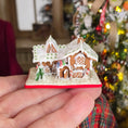 Load image into Gallery viewer, Miniature gingerbread house on a hand, held in front of a decorated Christmas tree.

