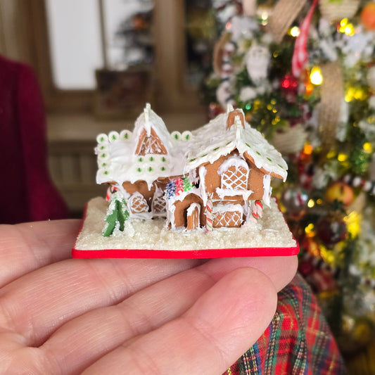 Miniature gingerbread house on a hand, held in front of a decorated Christmas tree.