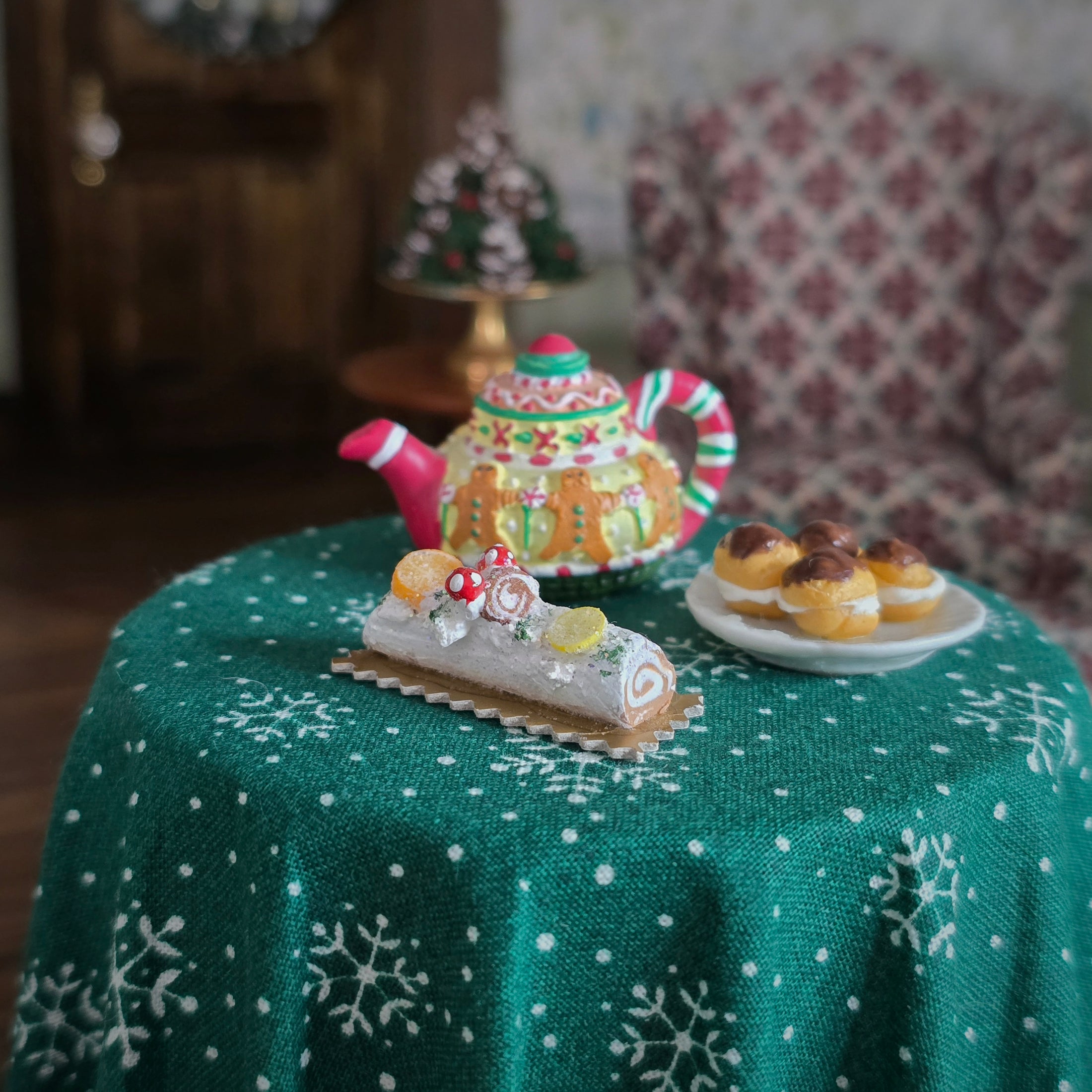 Miniature Christmas-themed teapot and pastries on a green tablecloth with snowflake pattern.