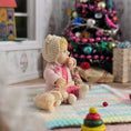 Load image into Gallery viewer, Catherine Muniere doll in a pink outfit with a knit hat sitting on a colorful blanket in front of a decorated Christmas tree.
