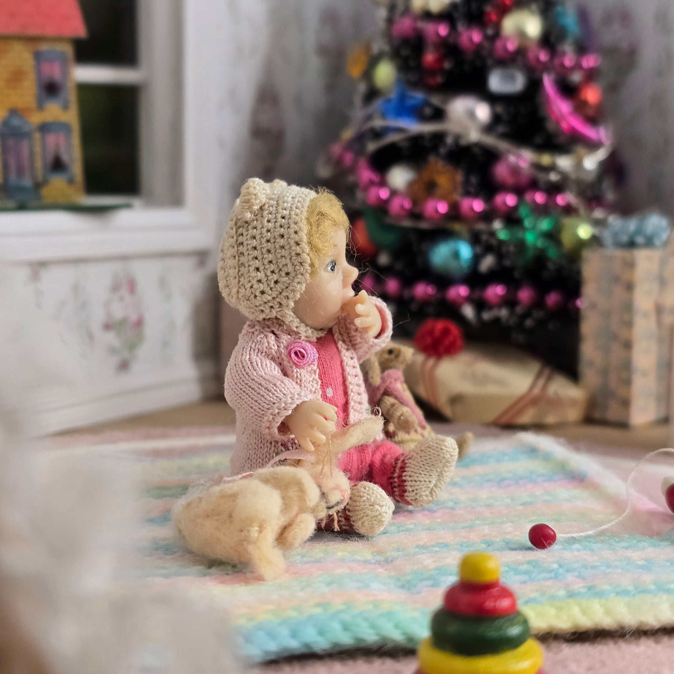 Catherine Muniere doll in a pink outfit with a knit hat sitting on a colorful blanket in front of a decorated Christmas tree.