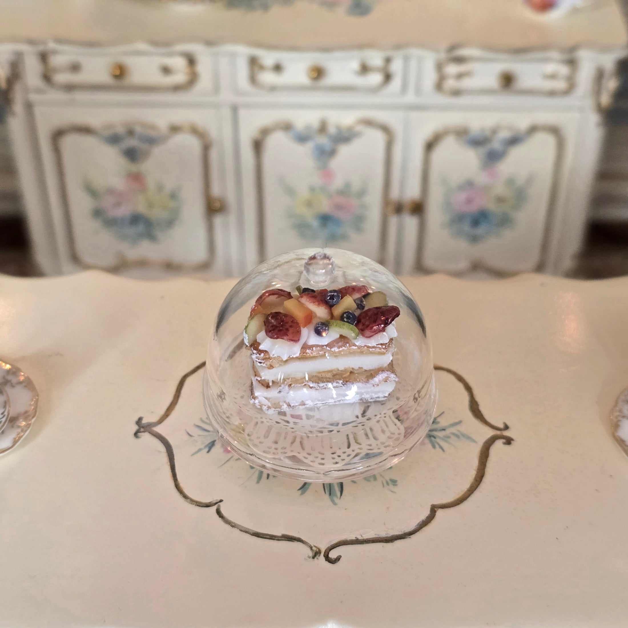 Miniature pastry in a dome on a decorative plate with a floral cabinet in the background