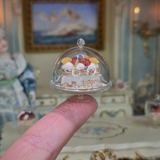 Miniature cake under a glass dome held by a finger with a decorative background