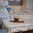 Load image into Gallery viewer, Cup of coffee and pastry on a marble surface with decorative elements
