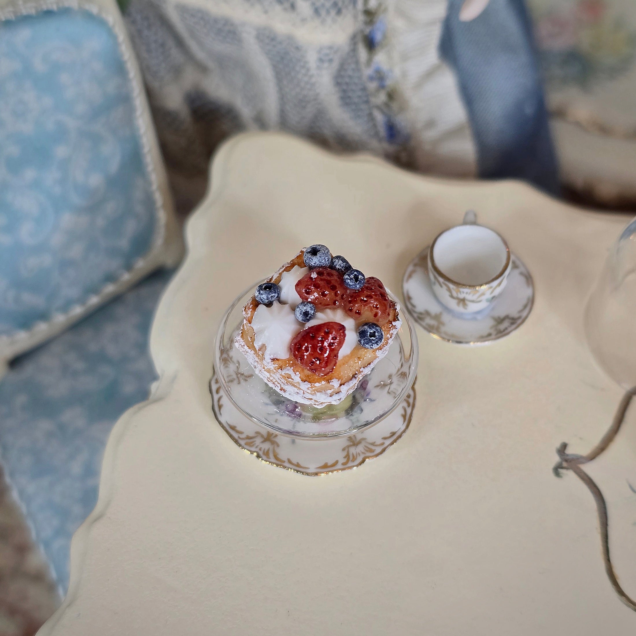 Small tart with berries on a decorative stand next to a teacup on a patterned surface.
