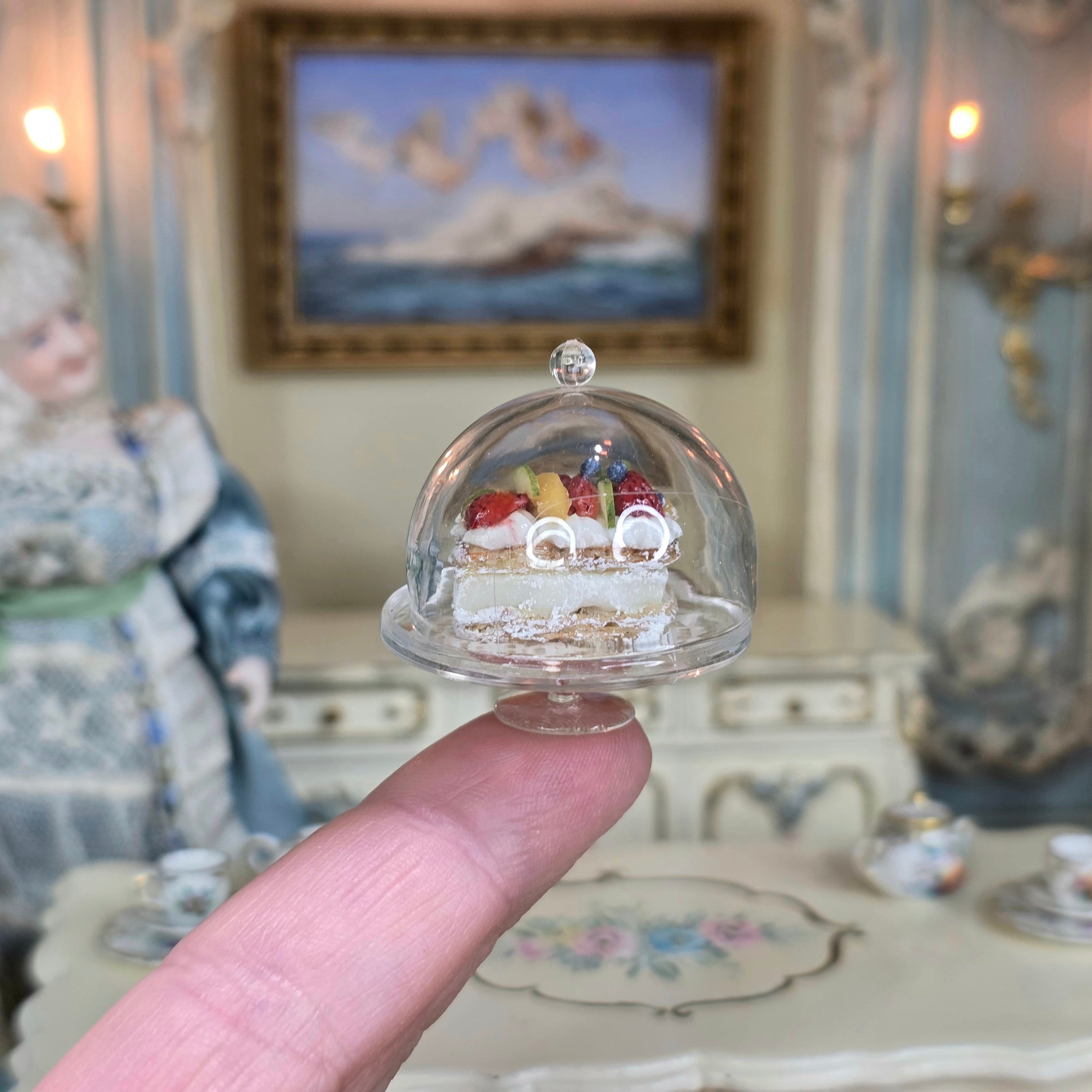 Miniature pastry under a clear dome held in front of a decorative background with a doll and furniture.