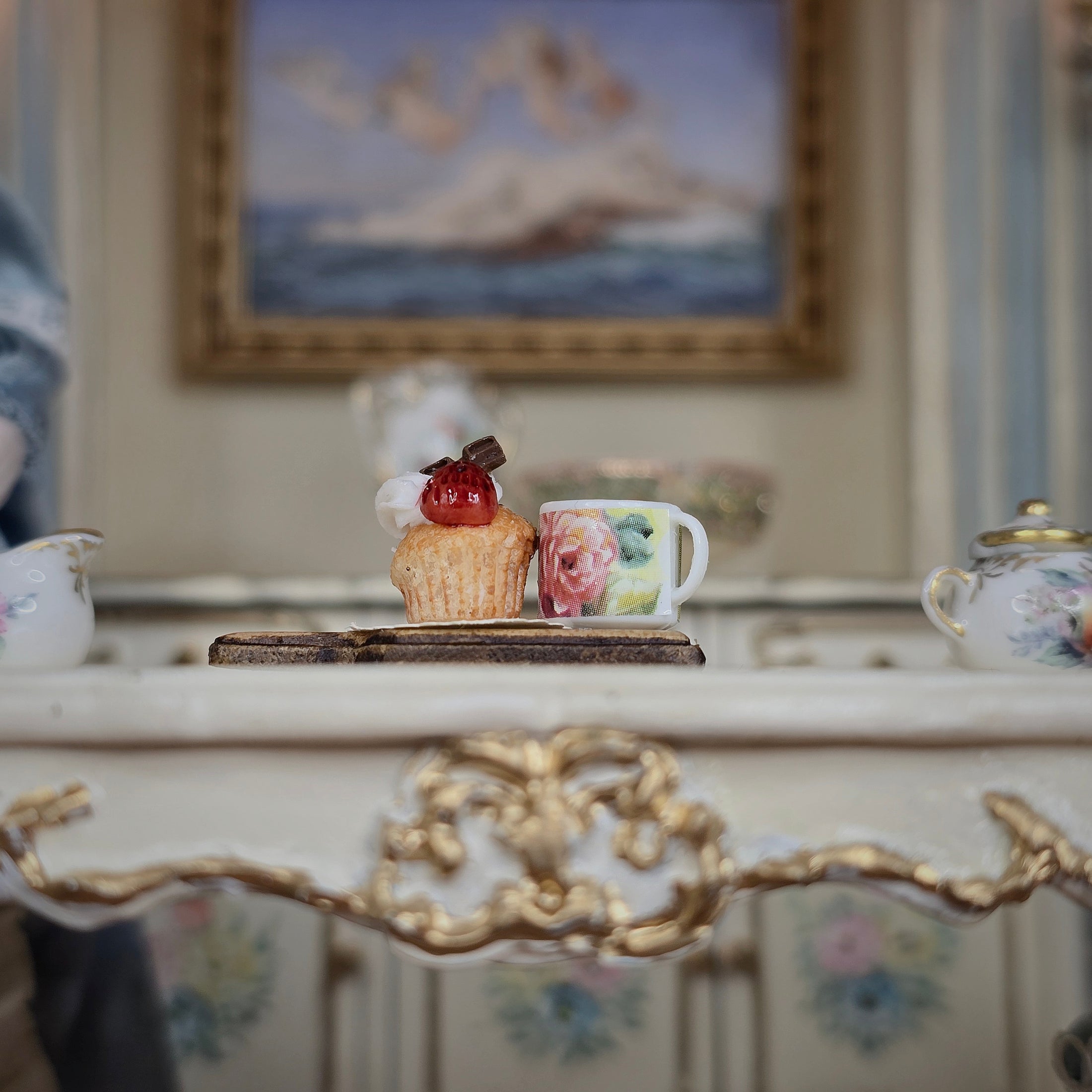 Decorative cupcake and mug on a ornate surface with a blurred painting in the background