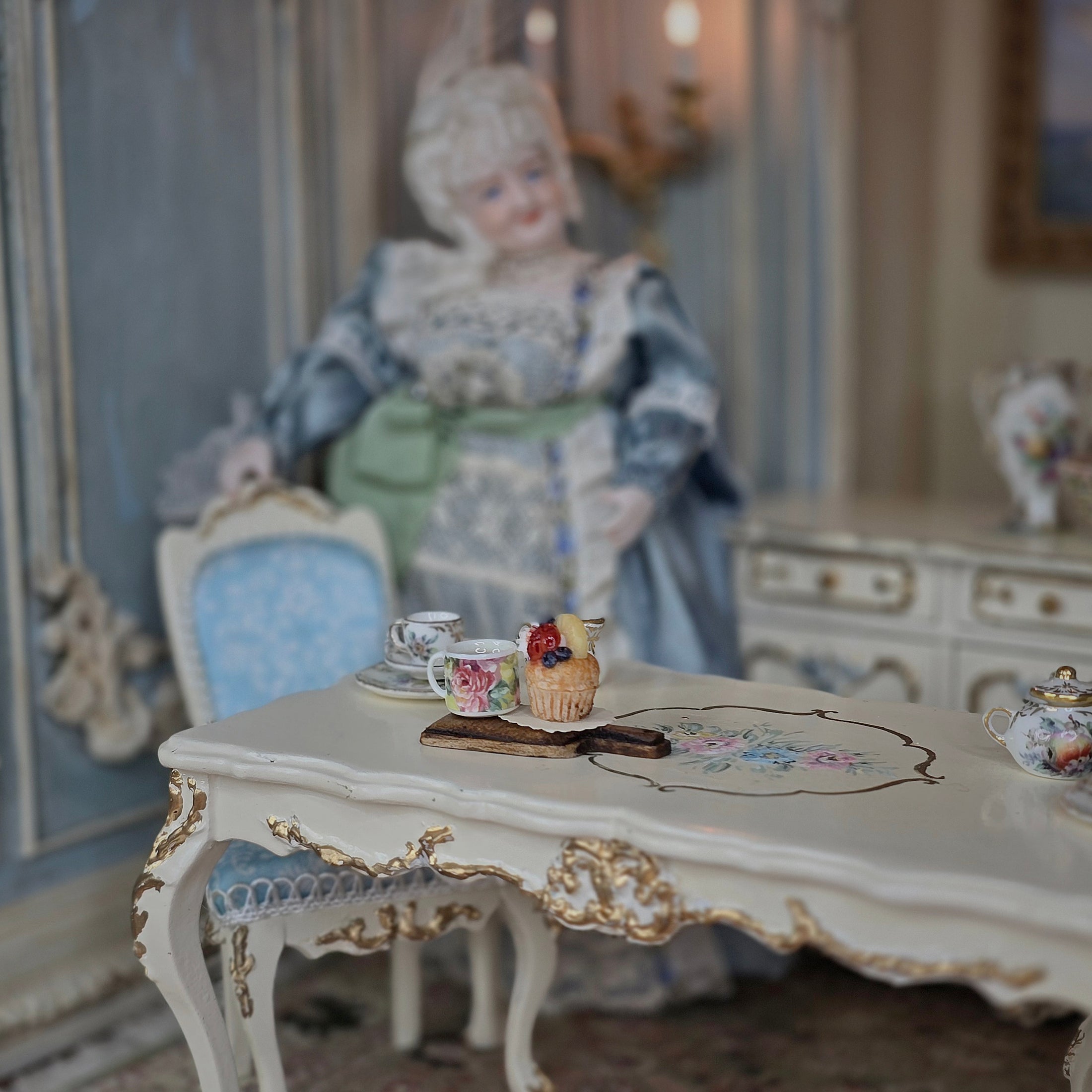 Miniature tea cup and cupcake diorama on a decorative table with a doll in the background
