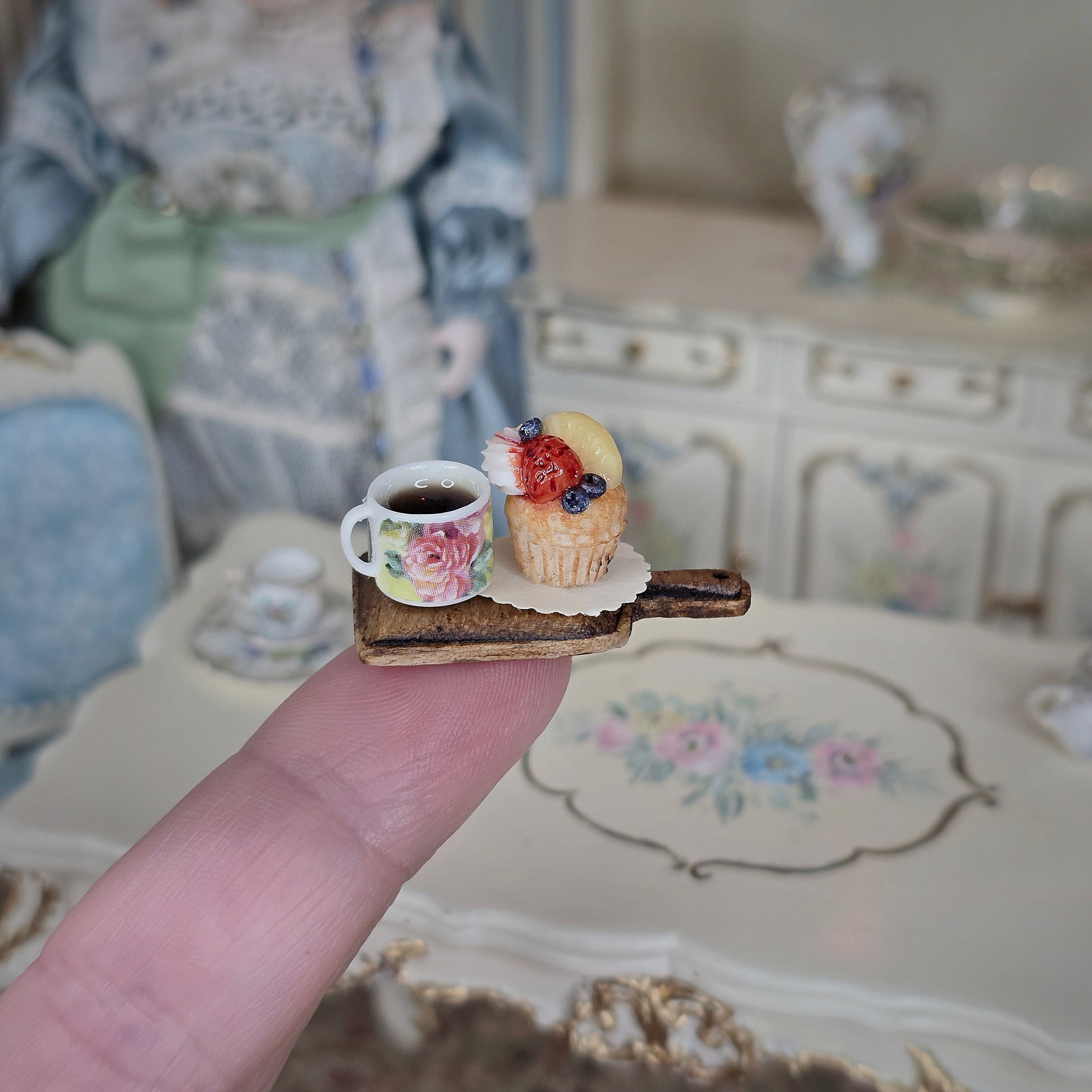 Miniature cup with floral design and a cupcake on a wooden tray held by a finger, with a blurred background of a room.