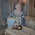 Load image into Gallery viewer, Vintage-style doll in a decorative setting with a table and chair.
